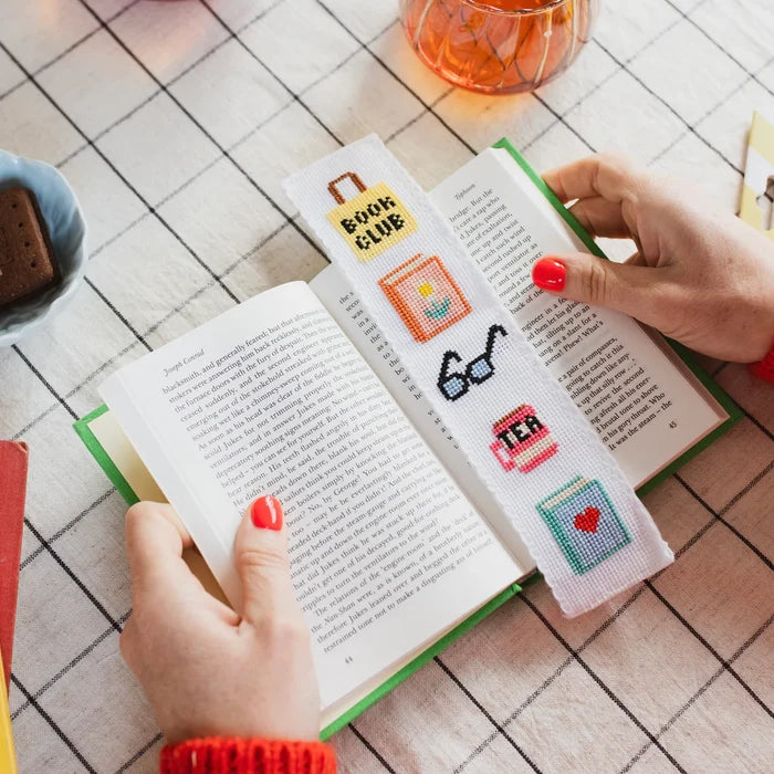 Person holding a book with a cross-stitched bookmark featuring various designs.