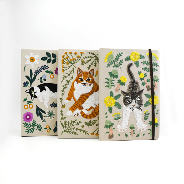 Three hardcover notebooks with linen covers featuring embroidered cats and wildflowers, arranged side by side on a white background.