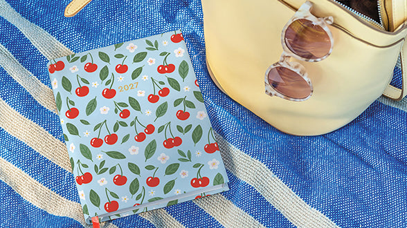 A light blue 2027 pocket calendar with a cherry and white flower pattern, styled on a blue and white striped blanket next to a cream leather bag and tortoiseshell sunglasses.