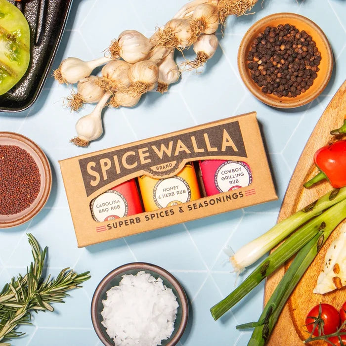 A Spicewalla brand spice and seasoning box displayed on a light blue surface with garlic, peppercorns, mustard seeds, salt, tomatoes, green onions, and rosemary arranged around it.