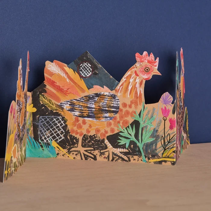 An angled, close-up view of a freestanding three-panel die-cut card featuring a stylized illustration of an orange speckled hen among garden plants.