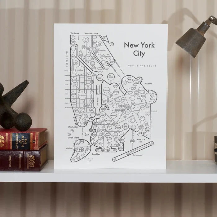 A black and white letterpress printed map of New York City displayed on a white shelf with a lamp and books in the background.