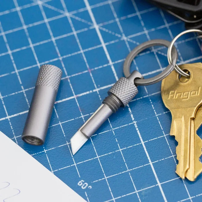 Metal keychain box cutter tool on a blue grid surface with a key and cylindrical object.