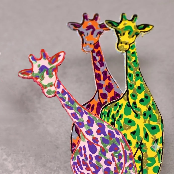 Three colorful giraffe bookmarks with long necks, decorated with vibrant hand-painted patterns in green, purple, orange, pink, and yellow, arranged against a neutral gray background.
