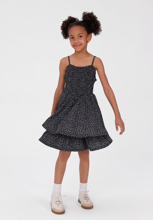 A young girl with curly hair is wearing a black and white micro-patterned spaghetti strap dress with a tiered skirt, paired with white socks and beige shoes, standing against a plain white studio background.