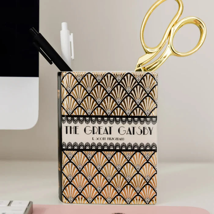 A rectangular vase shaped as a book featuring the Great Gatsby cover. It is shown holding a black pen, a white highlighter, and gold scissors on a desk.