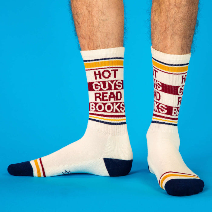 A pair of crew socks worn on a person's feet, featuring the text 'HOT GUYS READ BOOKS' in red, white, and navy blue stripes, set against a solid blue background.