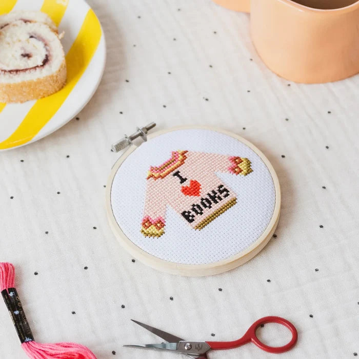 Cross-stitched hoop with 'I love books' sweater design on a polka dot tablecloth with sewing tools and a pastry.