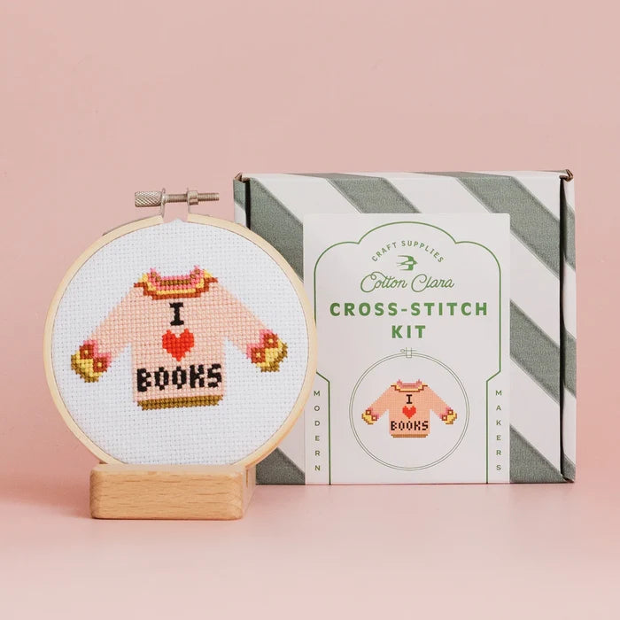 Cross-stitch kit with completed design of a sweater saying 'I love books' next to its packaging on a pink background.