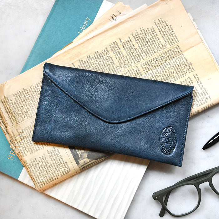 Blue leather long wallet featuring an oval NYPL book binding stamp, on top of books with glasses and a pen nearby