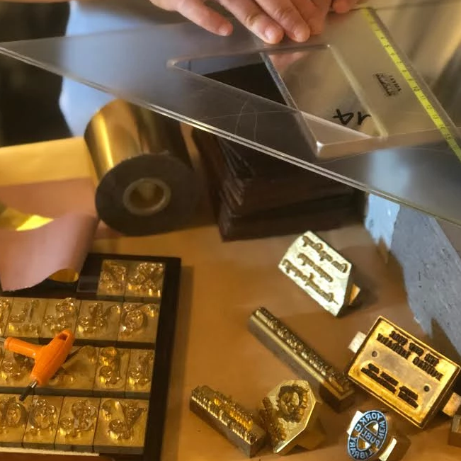 A close-up, overhead view of a bookbinder's workspace featuring brass type pieces, gold foil rolls, and a person using a large plastic set square to align materials.