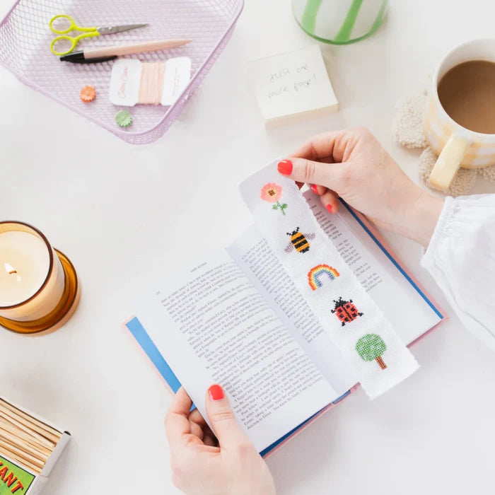 Person holding a book with a cross-stitched bookmark with a nature design that includes a tree, rainbow, bee, and flower on a desk with stationery items.