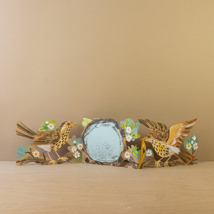 A three-panel freestanding die-cut card fully opened, showcasing two illustrated songbirds amidst leafy branches framing a central round space labeled "Message."