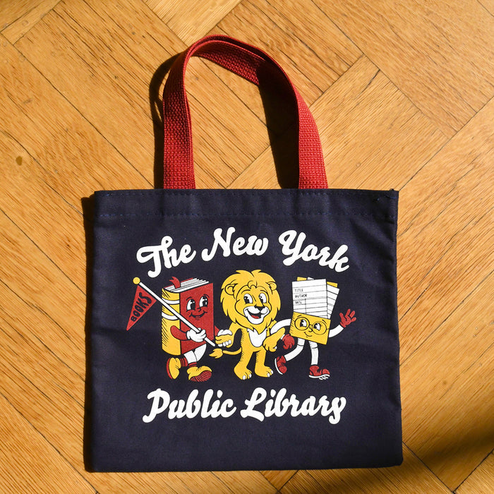 A navy blue canvas tote bag with red handles, featuring a colorful graphic of cartoon book and library characters with the text 'The New York Public Library' on a wooden floor background.