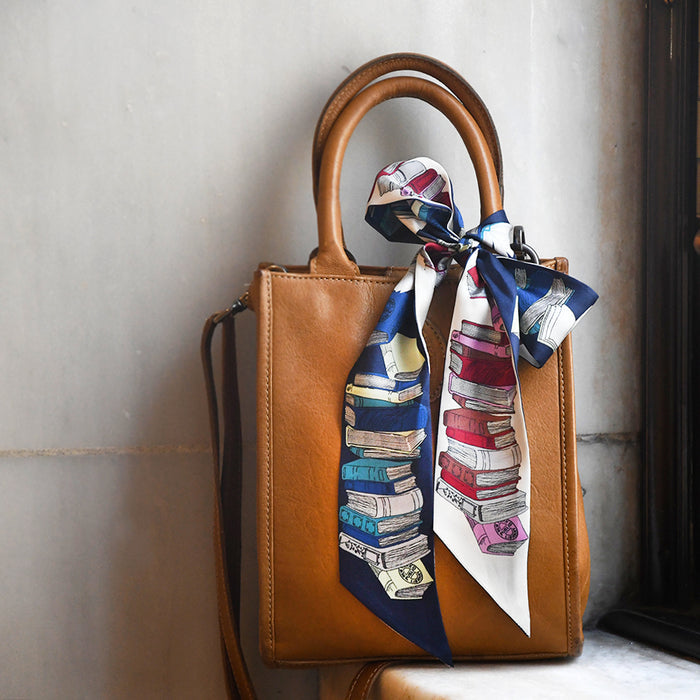 Brown handbag with a double-sided twilly scarf featuring illustrations of book stacks with an oval NYPL stamp. Twilly is tied around the handle against a neutral background.