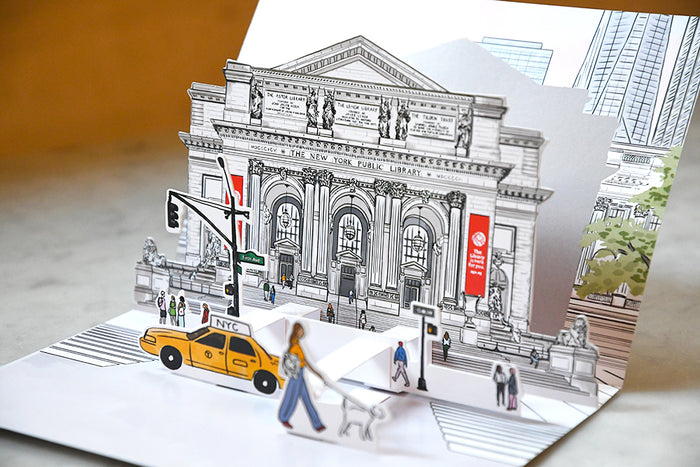 Pop-up  greeting card featuring a colorful, detailed sketch of the New York Public Library main entrance with a yellow taxi and pedestrians in the foreground, shown with a plain white envelope.