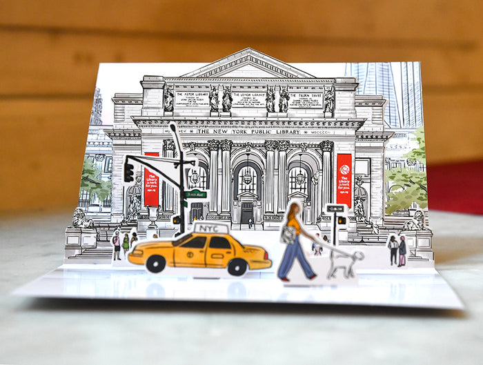 Pop-up greeting card featuring a colorful, detailed sketch of the New York Public Library main entrance with a yellow taxi and pedestrians in the foreground, shown with a plain white envelope.