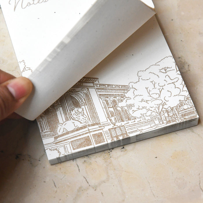 A close-up of a hand flipping through the notepad's pages, highlighting the paper's thickness and the textured Risograph library illustration at the bottom.
