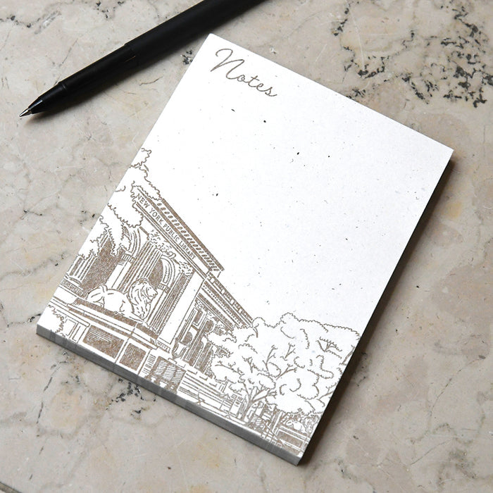 A top-down view of the full notepad with a black pen in the corner. It features a large blank writing area and a brown-ink illustration of the library across the bottom.
