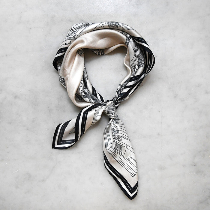 A black and white silk scarf with an architectural print, knotted loosely and displayed on a white marble surface.