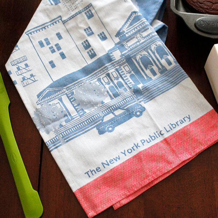 An off-blue cotton tea towel with a darker blue illustration of The New York Public Library's Stephen A. Schwarzman Building and the city's skyline. Red detailing at the bottom.