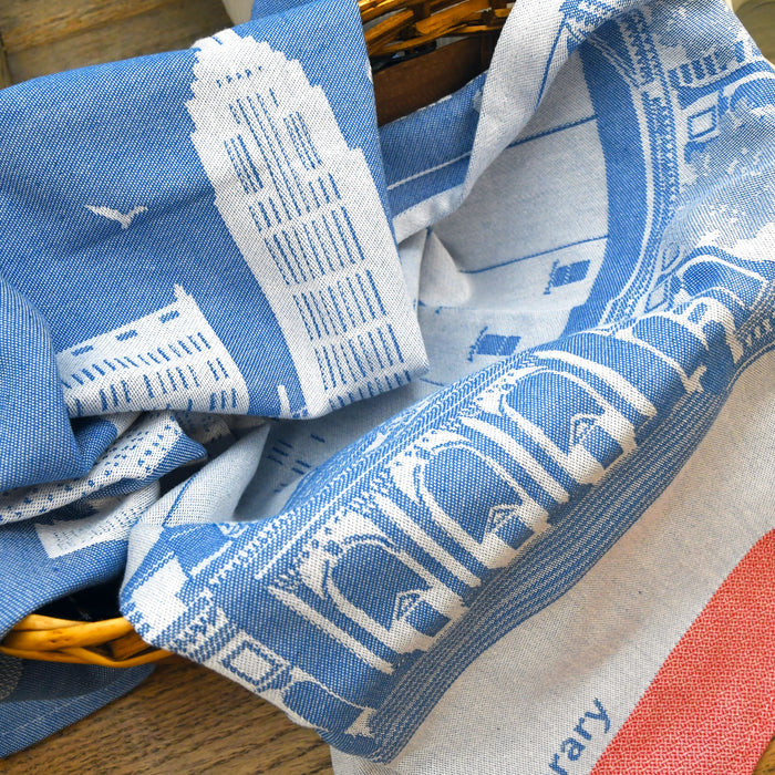 An off-blue cotton tea towel with a darker blue illustration of The New York Public Library's Stephen A. Schwarzman Building and the city's skyline. Red detailing at the bottom.