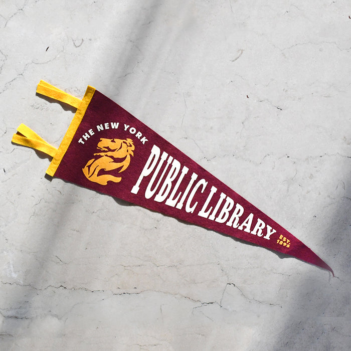 A triangular maroon and gold pennant flag for The New York Public Library, featuring a gold lion logo and white lettering, laid diagonally on a light gray stone surface.