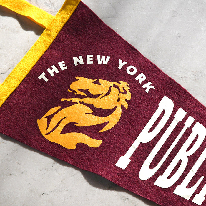 A close-up of a felt pennant flag with a yellow handle, featuring the text 'THE NEW YORK PUBLIC' and a yellow lion graphic on a maroon background, placed on a light gray textured surface.