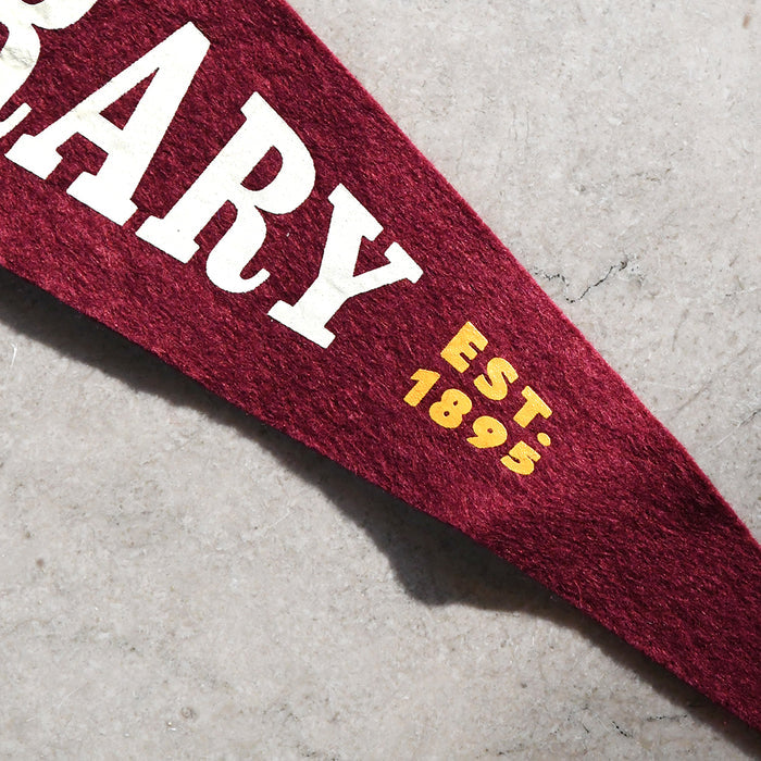 A close-up of a maroon felt pennant banner with white text reading 'MARY' and yellow text reading 'EST. 1895', laid diagonally on a light gray textured surface.
