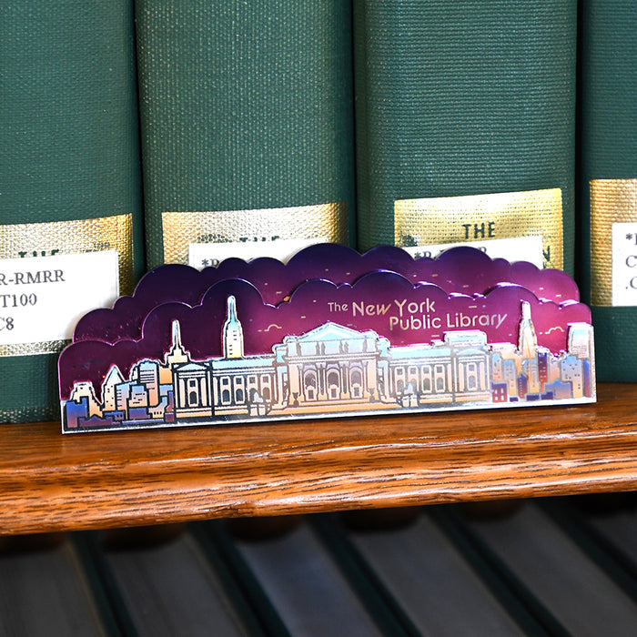 An acrylic magnet shaped like NYPL's Stephen A. Schwarzman Building with the skyline in the background with a purple cloud background, displayed on a wooden bookshelf in front of green book spines