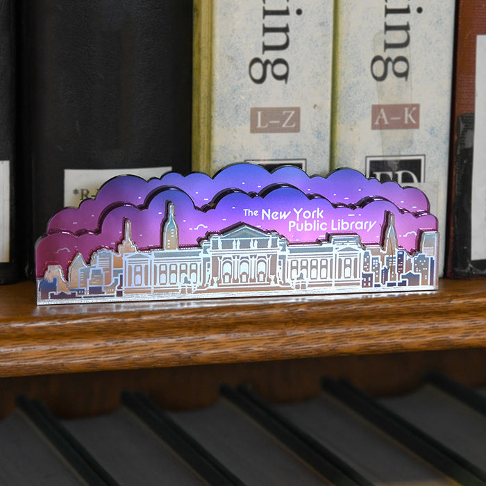 An acrylic magnet shaped like The New York Public Library building skyline, with a purple-to-pink iridescent gradient sky and white building outline, displayed on a wooden shelf in front of books.