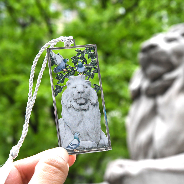 A hand holding a bookmark featuring an image of a marble lion with a bird perched on its head, with a blurred background of a similar statue.