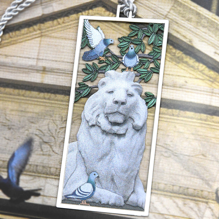 Close-up of the metal bookmark featuring one of the NYPL lions during summer with pigeons and green trees.