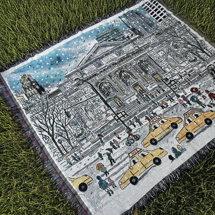 A throw blanket with a printed design featuring an illustration of NYPL's flagship building during the winter season on grass.