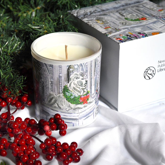 Decorative candle with a festive NYPL lion design in a box, surrounded by red berries and greenery.
