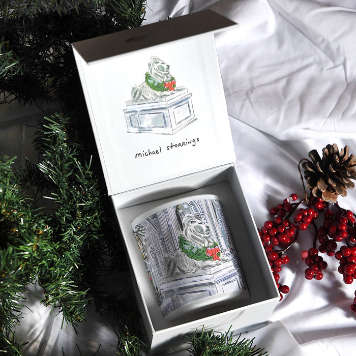 Decorative candle with a festive NYPL lion design in a box, surrounded by red berries and greenery.