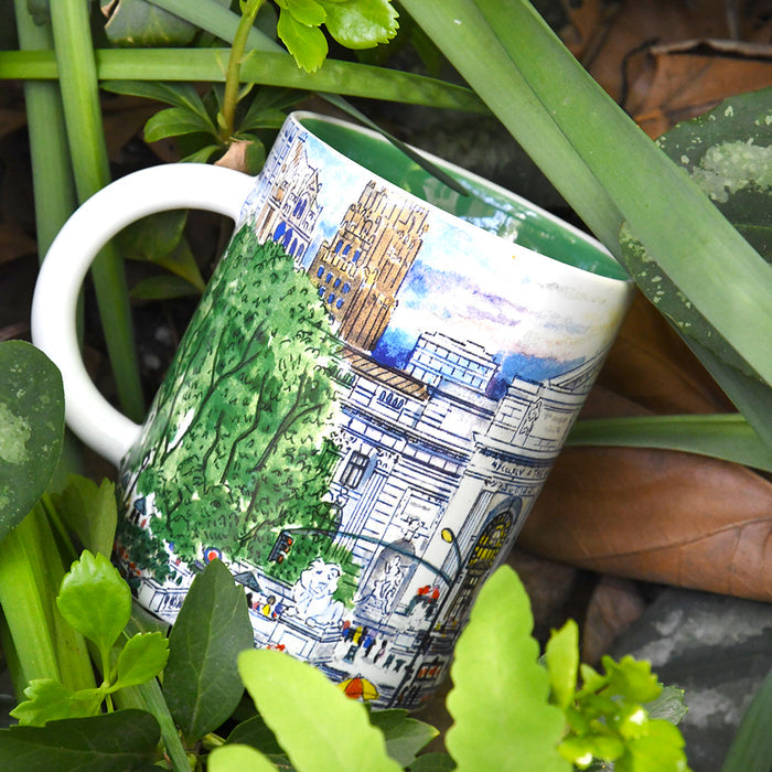A white ceramic mug with a hand-painted illustration of the facade of NYPL's flagship building during spring season, nestled among green leaves and plants.