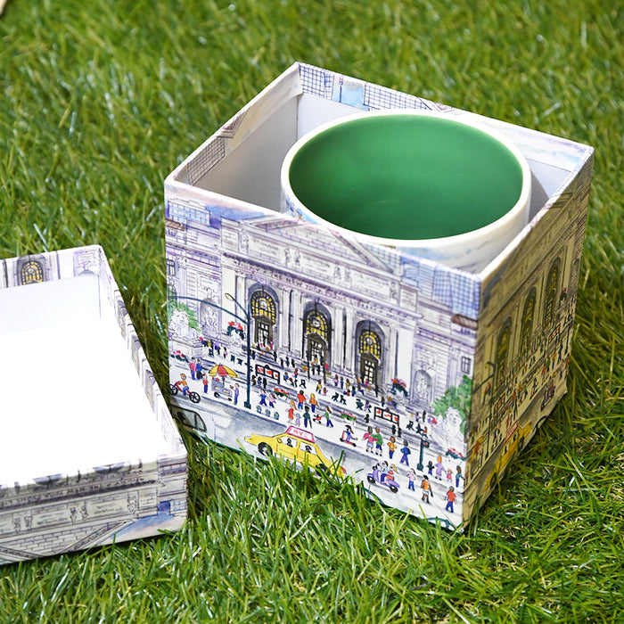A square ceramic mug with a green interior sits inside a decorative printed box featuring the NYPL's flagship building during spring season, placed on green grass. The matching lid rests to the left.