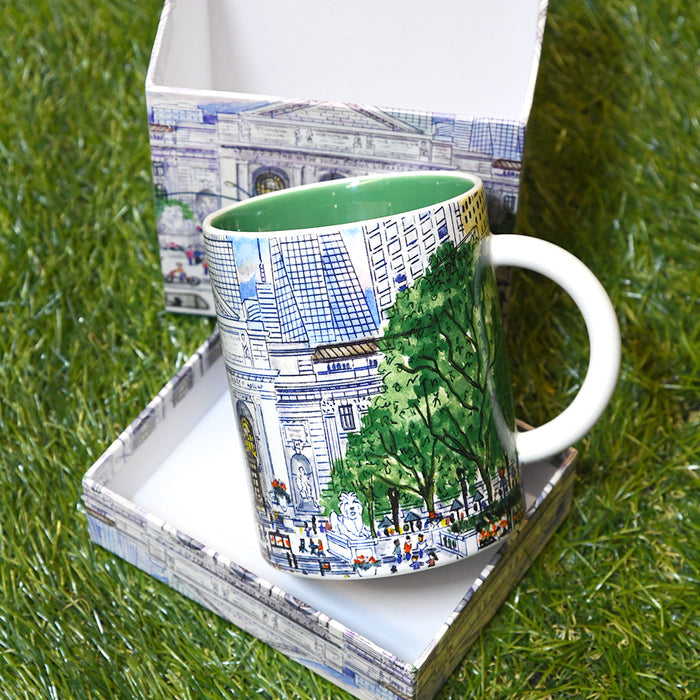 A white ceramic coffee mug with a colorful illustration of the NYPL's flagship building during spring season, placed on top of its open printed box on green grass.