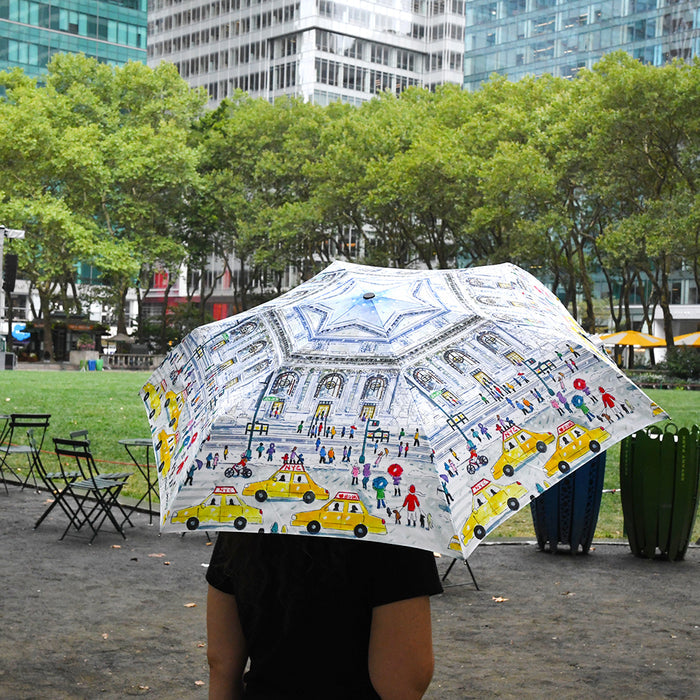 Person holding a colorful umbrella featuring an illustration of NYPL's flagship building during the winter season  in an urban park setting