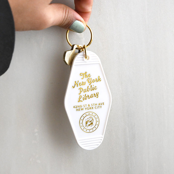 White motel-style keychain featuring text "The New York Public Library" and its 42nd Street address embossed in shiny gold lettering, along with the library's lion seal.