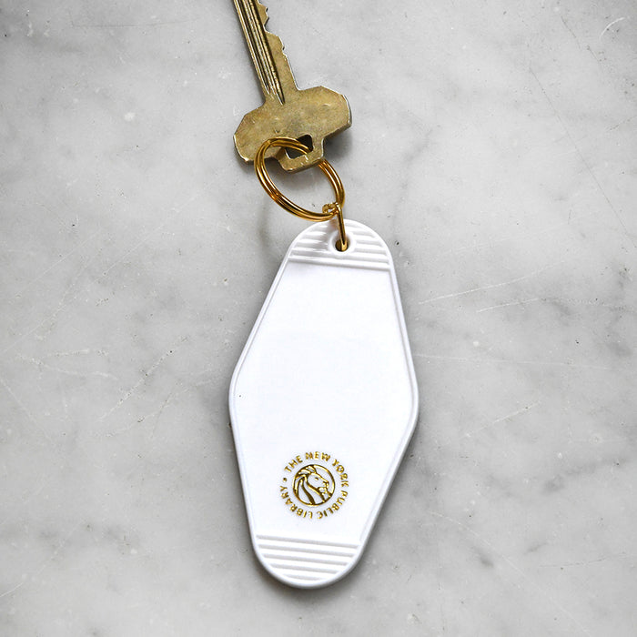 The back view of white plastic motel-style keychain on marble surface. This side is mostly blank, except for a small, circular gold logo near the bottom featuring a lion's head surrounded by the words "The New York Public Library."