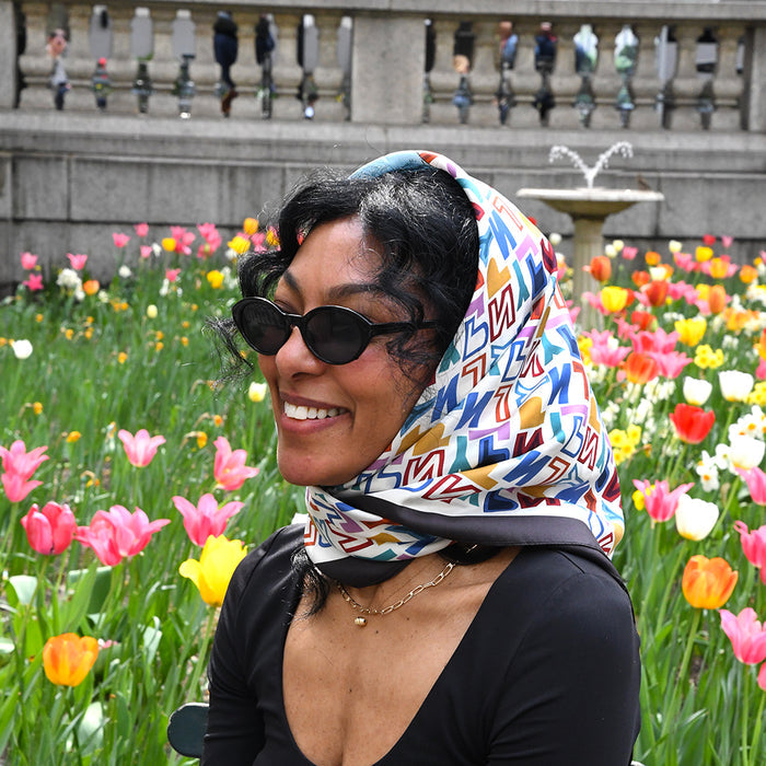 Woman with a headscarf featuring a colorful 'NYPL' pattern and sunglasses standing in a garden with tulips.