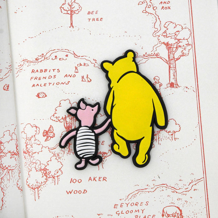 A rubber magnet shaped as Winnie-the-Pooh and Piglet from the Hundred Acre Wood, placed on a book with a red line drawing background.