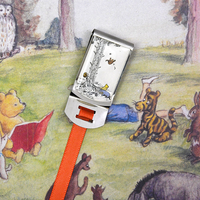 A silver clip bookmark with an orange ribbon. The clip features a classic sketch of Christopher Robin, Pooh, and Eeyore looking up at Tigger in a tree.