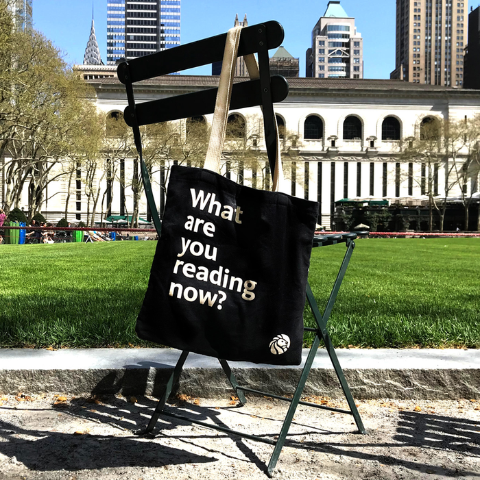 A black cotton tote bag with off-white handles featuring  the quote 'What are you reading now?'  displayed on a metal chair with a library and cityscape in the background.