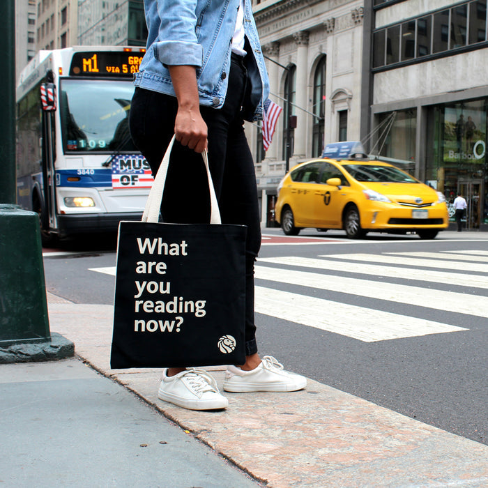 A black cotton tote bag with off-white handles featuring  the quote 'What are you reading now?'.