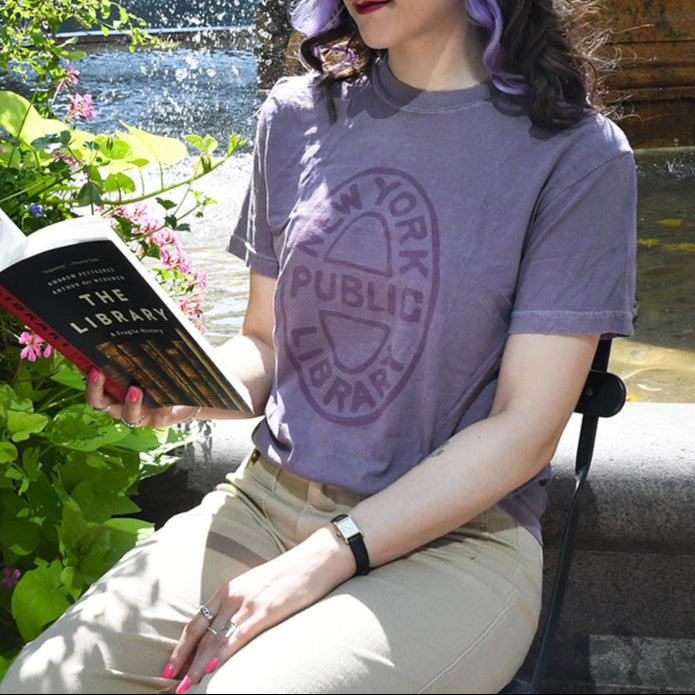 Person wearing a New York Public Library purple shirt that features an oval NYPL bookbinding stamp, sitting by a body of water with flowers.