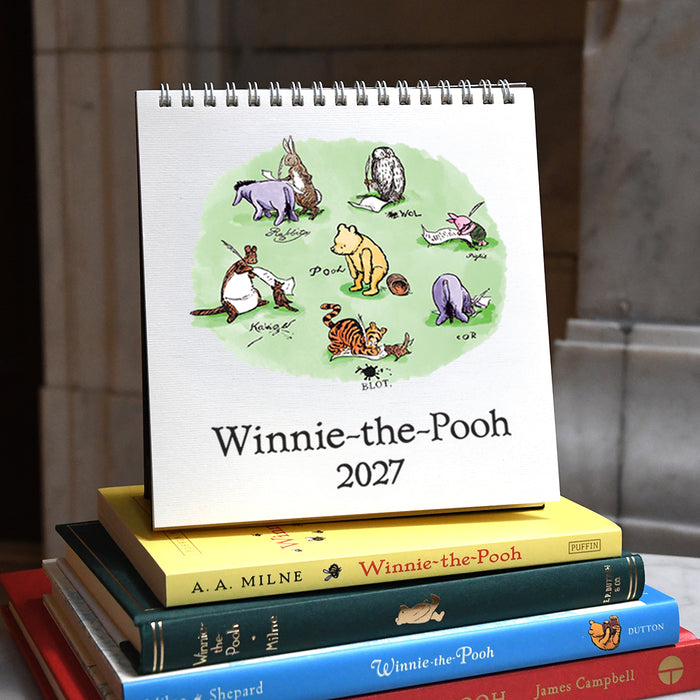 A spiral-bound 2027 Winnie-the-Pooh calendar displayed upright on top of a stack of Winnie-the-Pooh books, with the calendar cover showing illustrated characters on a light green circle and the title 'Winnie-the-Pooh 2027'.