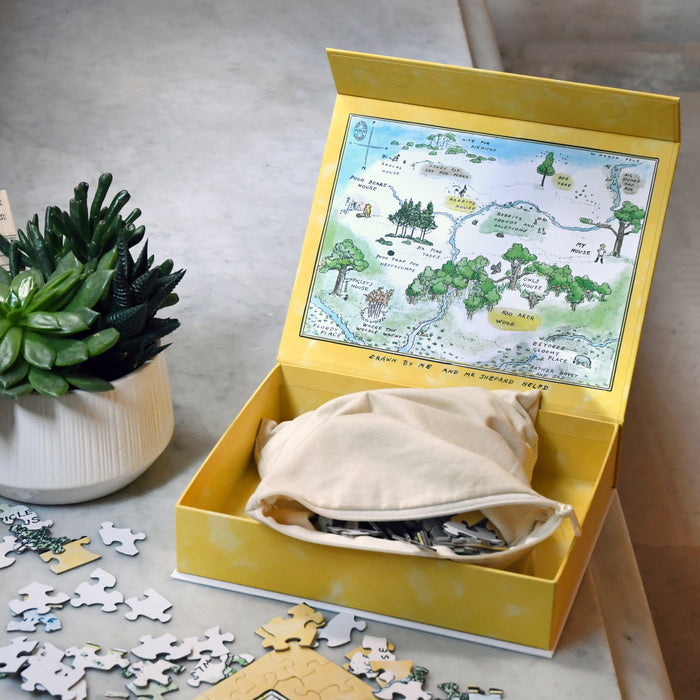 An open yellow gift box containing a zippered cloth bag of puzzle pieces, with the inside lid featuring a colorful "100 Aker Wood" map from Winnie-the-Pooh.
